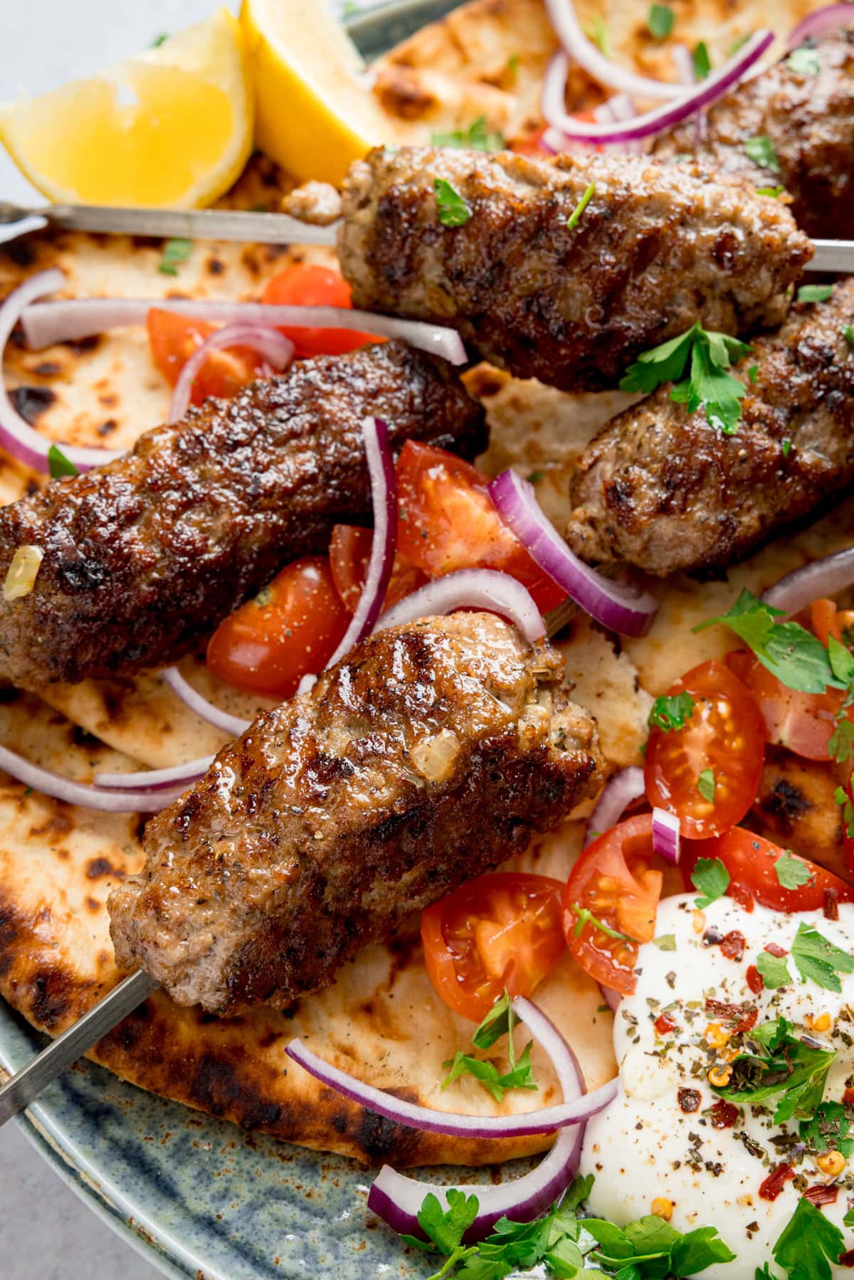 Tall close-up image of grilled lamb kofta skewers on flatbreads, on a plate, with sliced tomatoes, red onions, lemon wedges, fresh herbs, and a side of whipped feta.