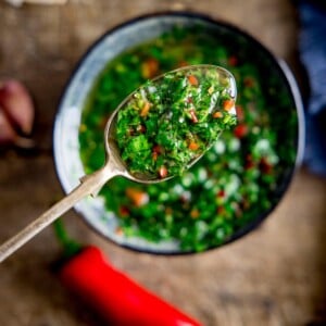 A spoon full of chimichurri sauve being shown woth a bowl full of chimichurri in the background out of focus.