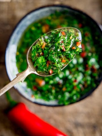 A spoon full of chimichurri sauve being shown woth a bowl full of chimichurri in the background out of focus.