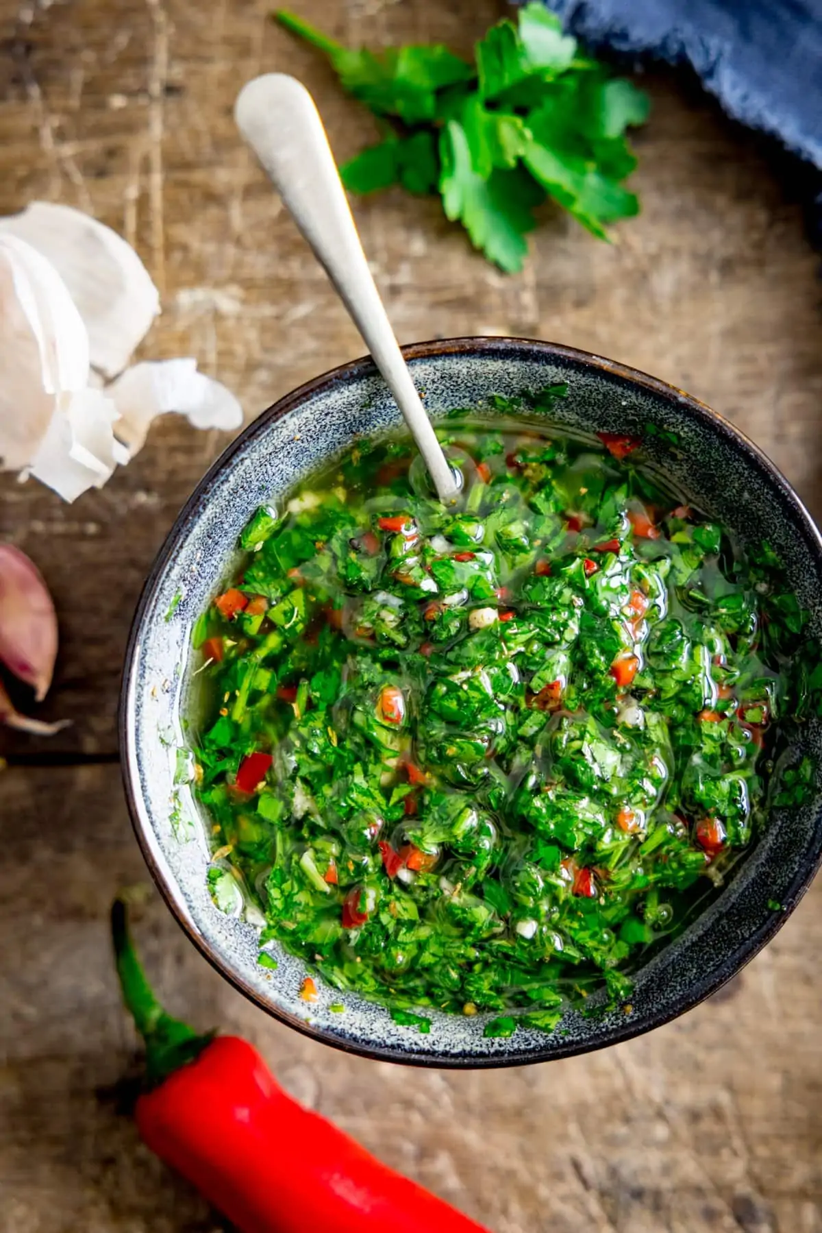 Overhead picture of a bowl full of chimichurri with a spoon sticking out, sat on a wooden background.