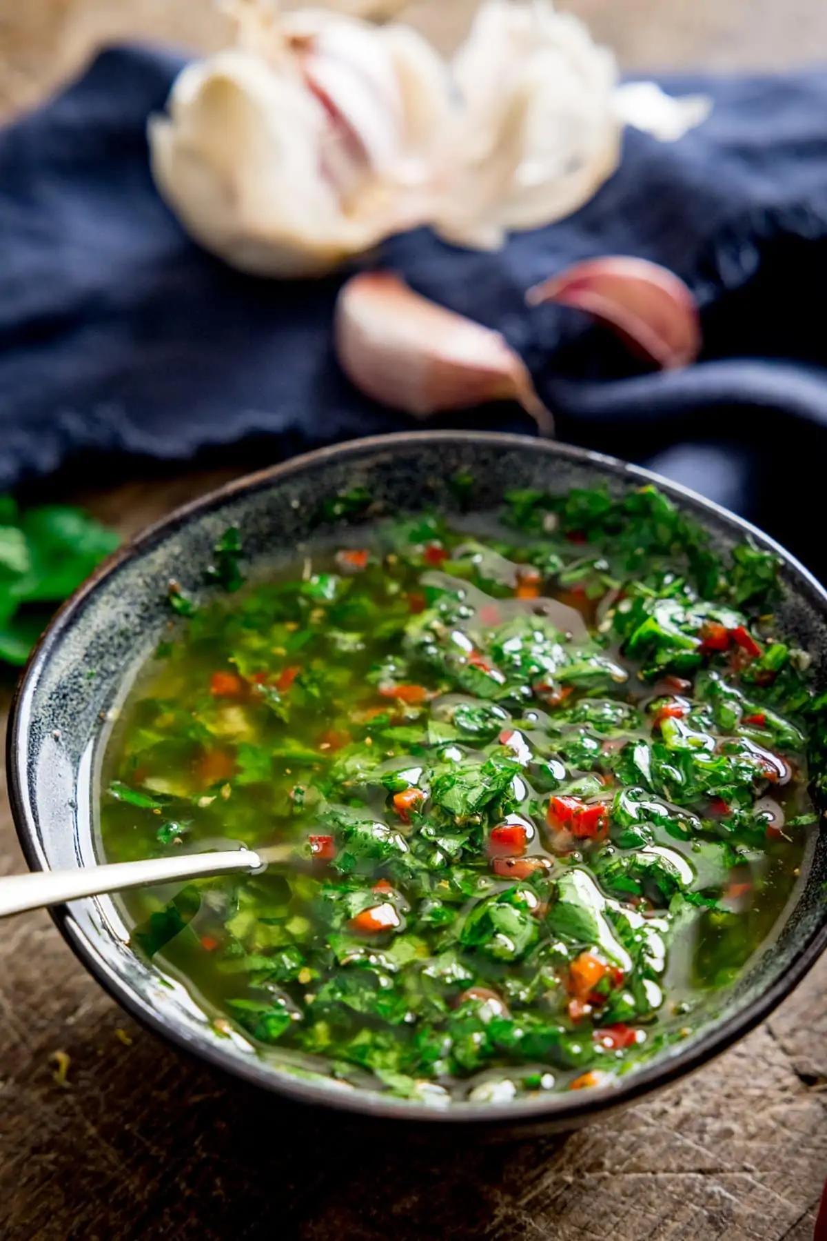 Dark bowl with Chimichurri sauce in and a blue napkin and garlic cloves out of focus in the background.