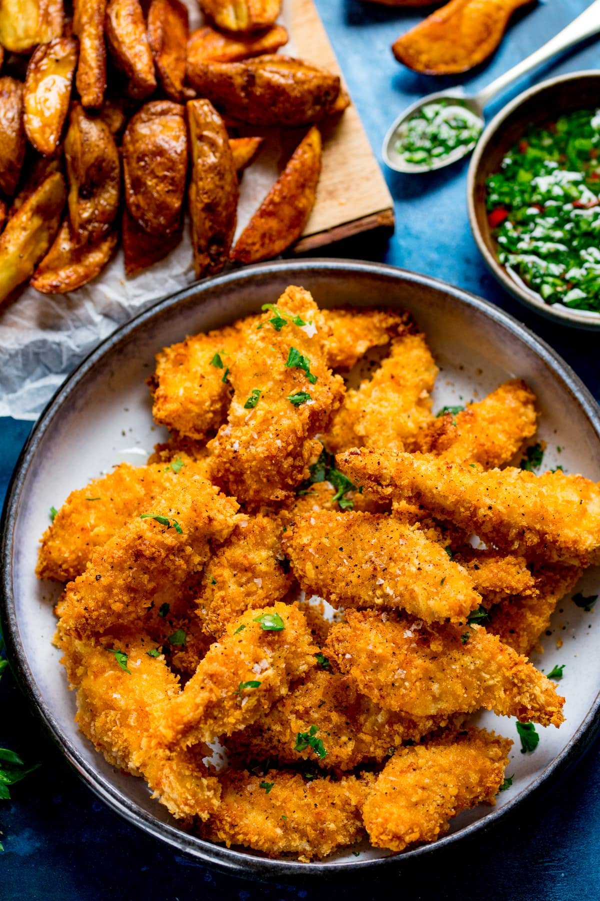 A bowl full of crispy baked chicken goujons with some potato wedges and chimichurri in the background.