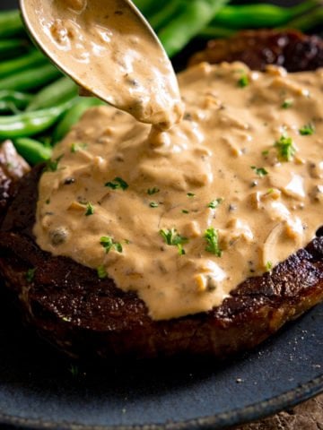 A cooked steak with peppercorn sauce being poured over the top with a spoon, green beans in the background on a blue plate