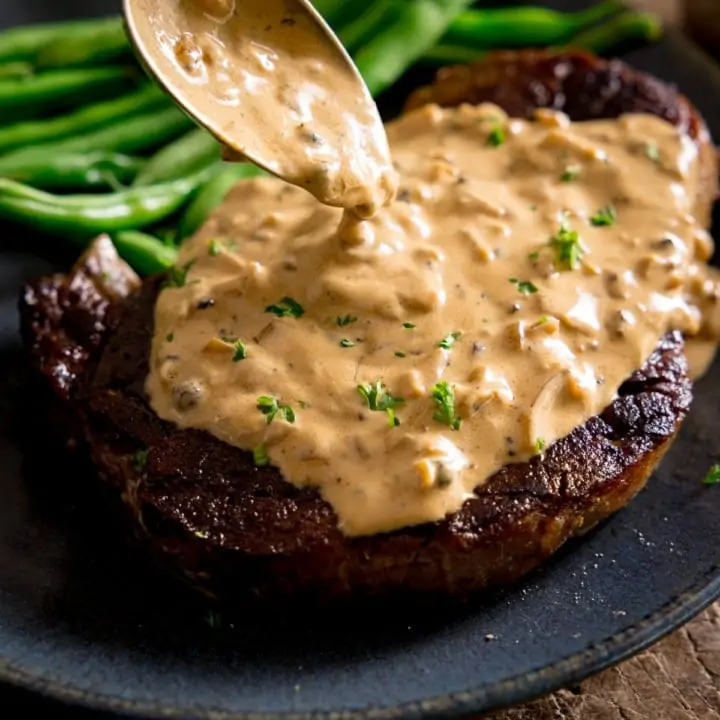 A cooked steak with peppercorn sauce being poured over the top with a spoon, green beans in the background on a blue plate