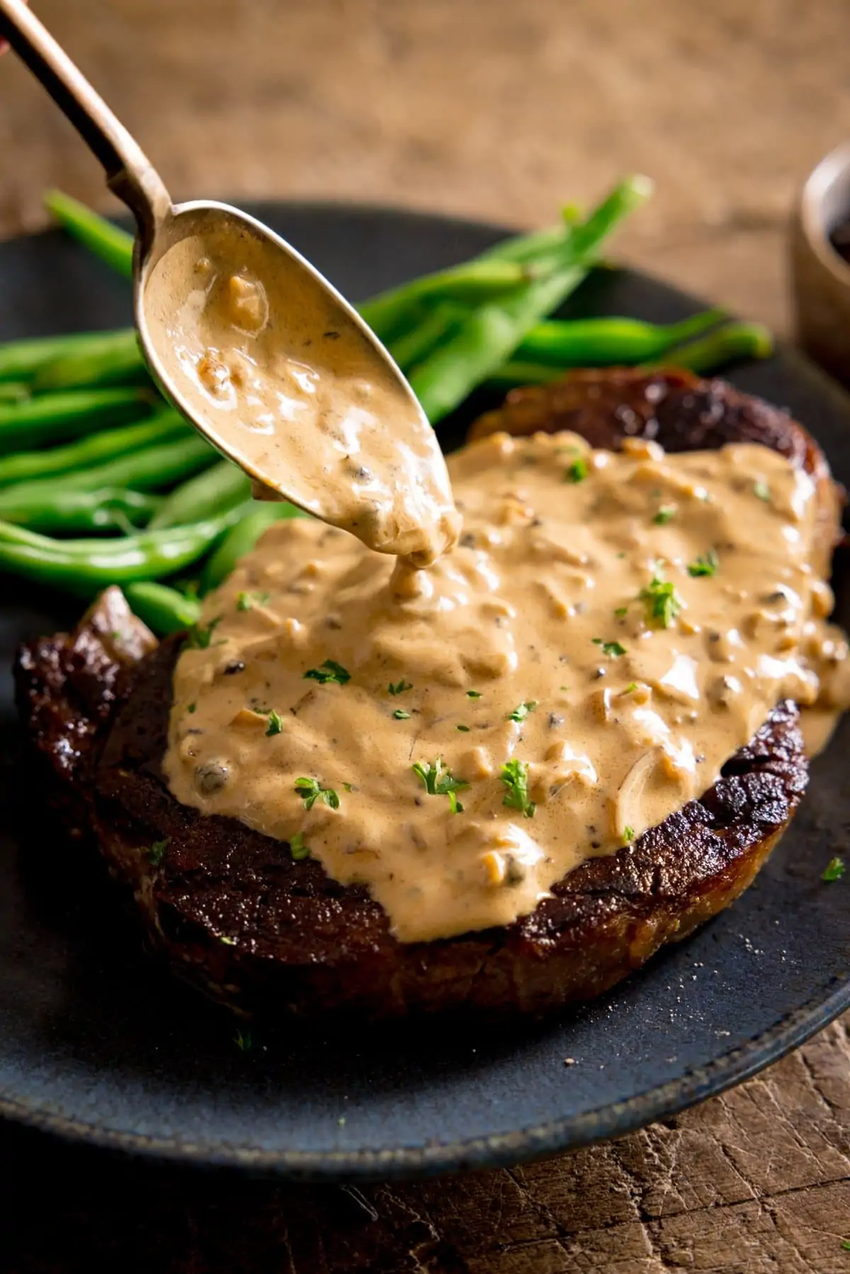 Peppercorn sauce being spooned over a cooked steak on a blue plate with green beans
