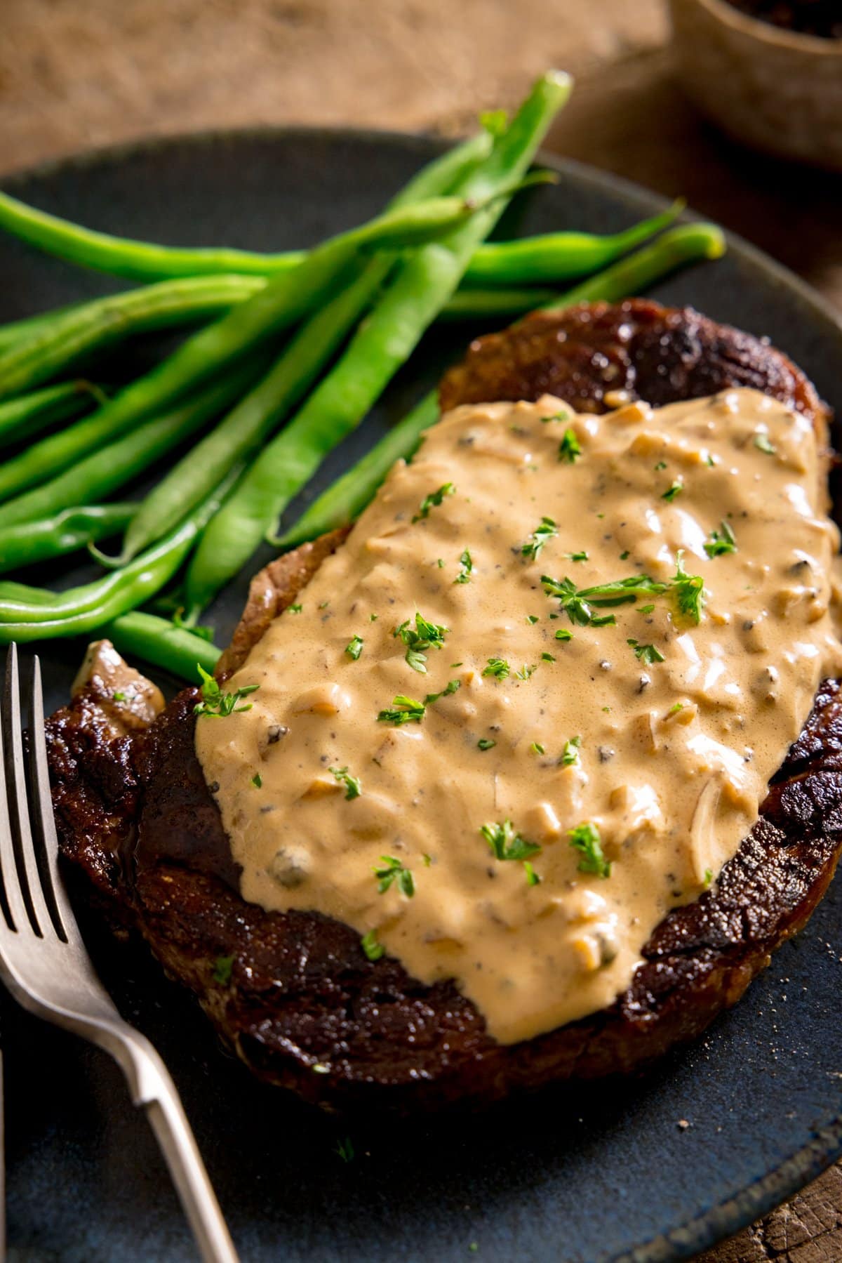 A tall image of a cooked steak with peppercorn sauce, served with green beans on a blue plate. There is a fork on the plate.