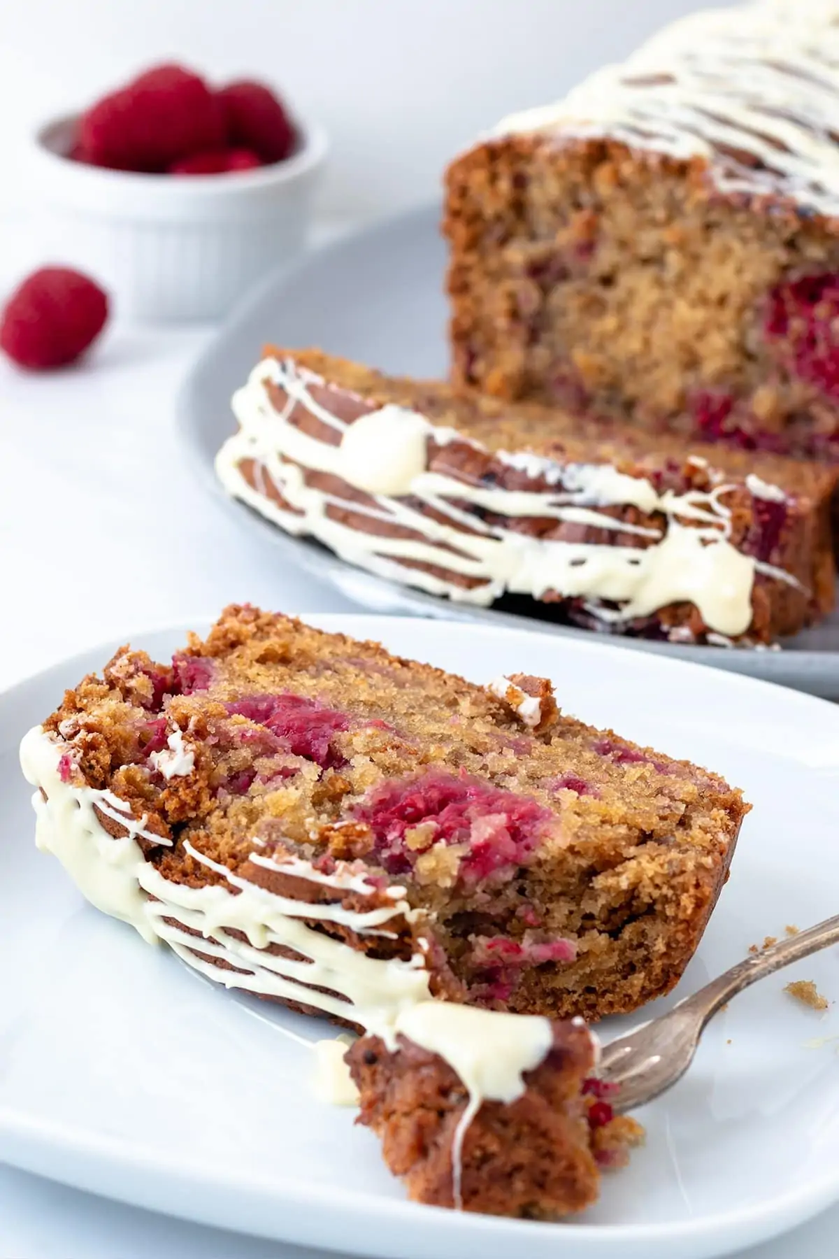 Close up image of a raspberry and white chocolate cake sat on a white plate showing the texture of the cake.  There is the rest of the cake in the background out of focus.