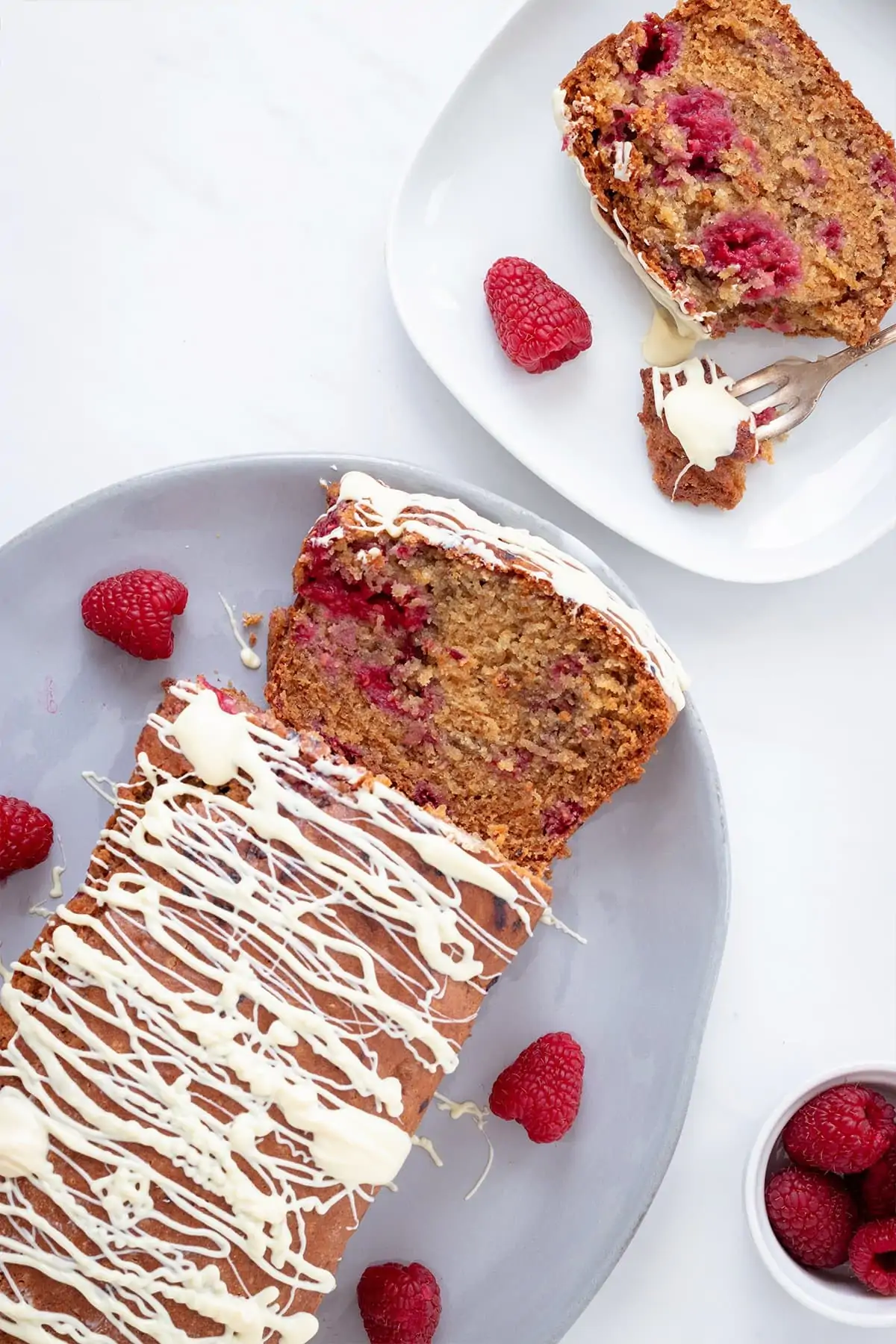 overhead image showing a raspberry cake drizzled with melted white chocolate on a white background with a bowl of fresh raspberries in the corner.