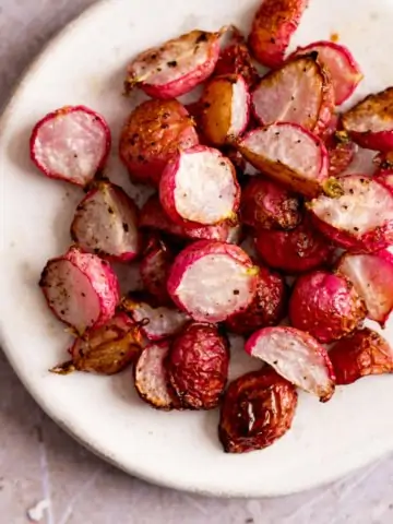 Square image of halved roasted radishes on a white plate, on a light background.