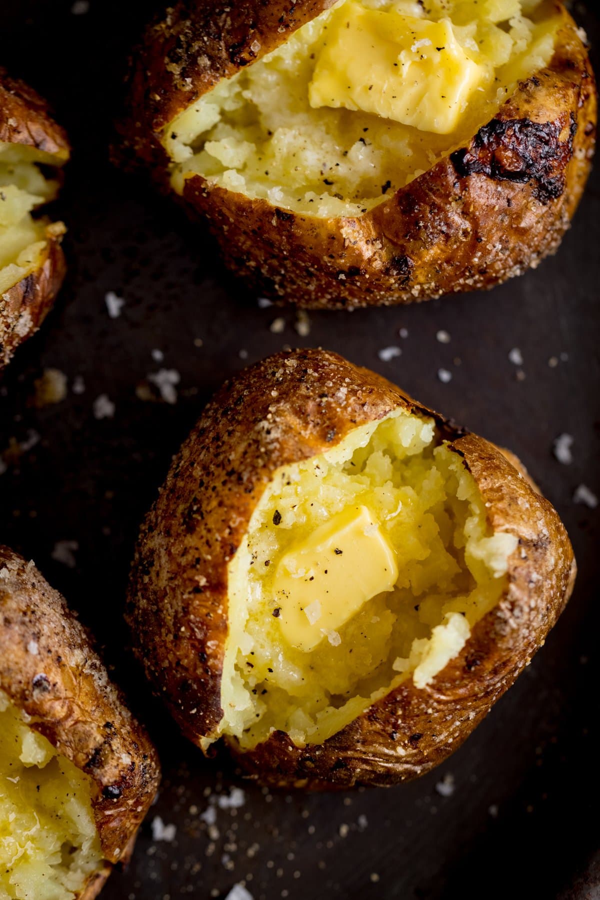 Overhead of an open, butter-filled baked potato on a dark surface. Further baked potatoes surround the main one.