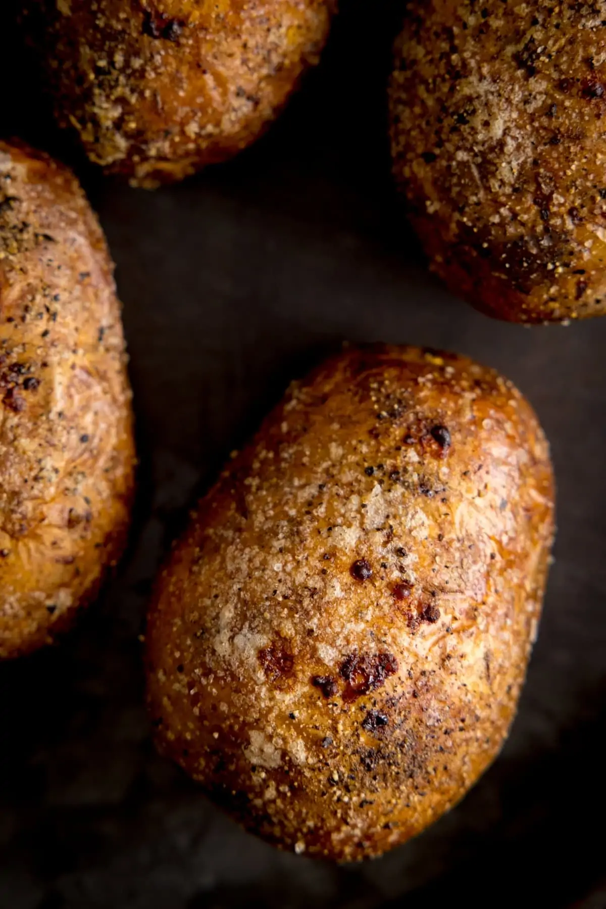 Overhead of an unopened baked potato on a dark tray. Further baked potatoes are arranged around the main one.