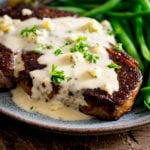 A cooked steak with blue cheese sauce over the top with some green beans, served on a blue plate