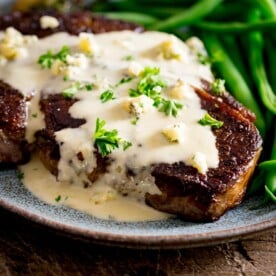 A cooked steak with blue cheese sauce over the top with some green beans, served on a blue plate