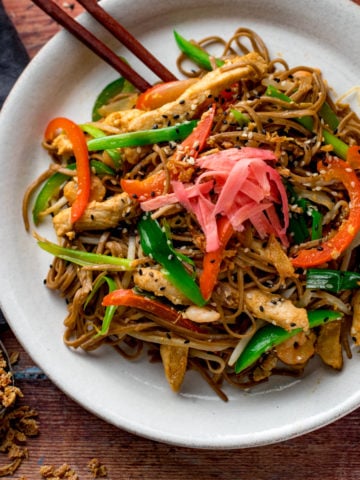 a square image of chicken yakisoba on a white plate with some chopsticks on the side, placed on a wooden board with some ingredients in the background