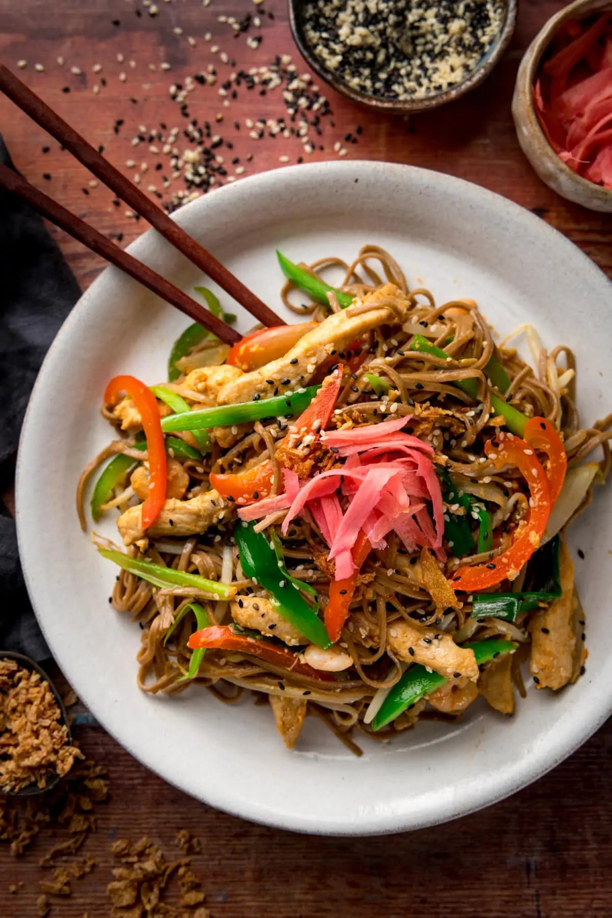 A tall image of chicken yakisoba on a white plate with some chopsticks, on a wooden board
