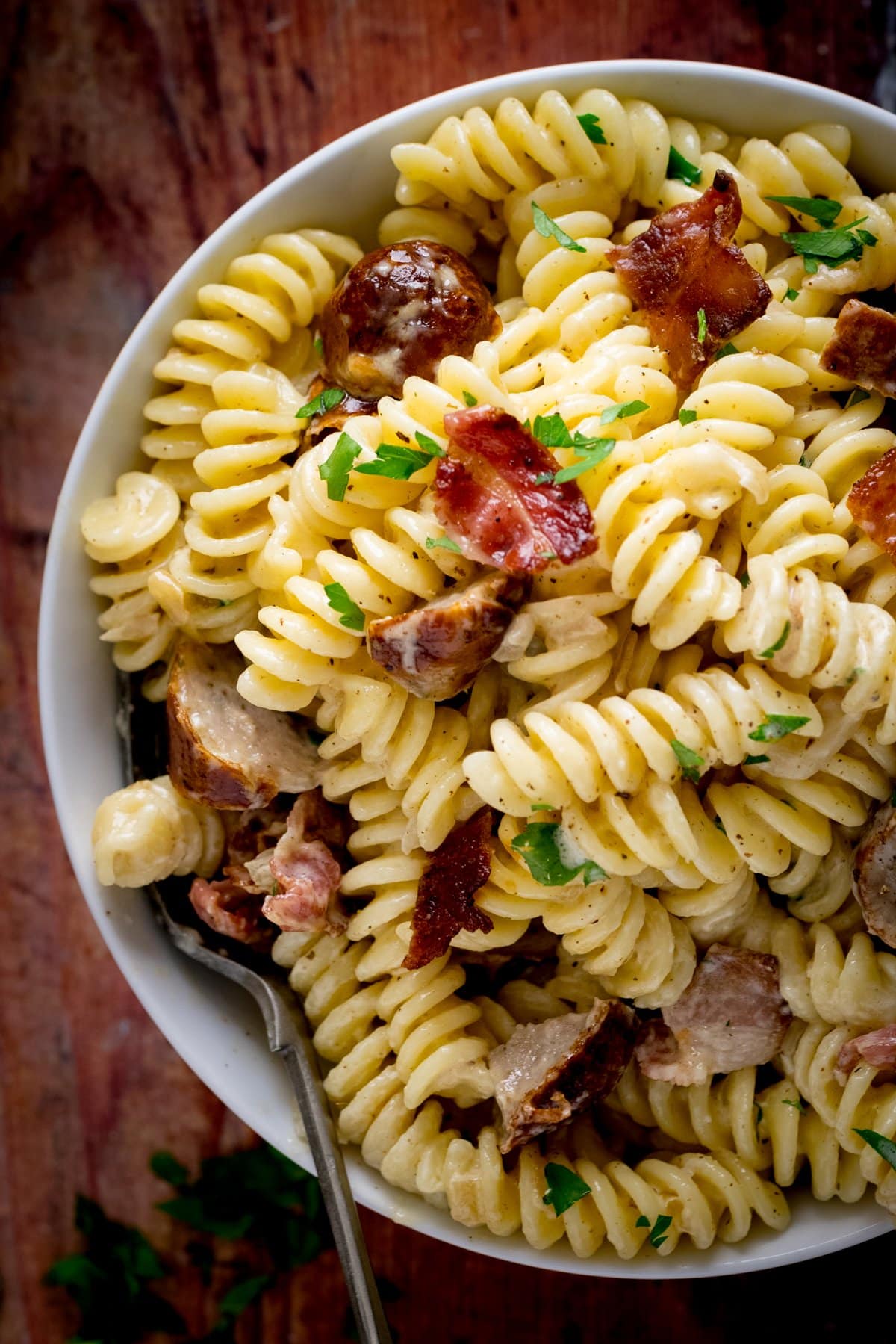 creamy pasta with sausage and bacon topped with parsley in a white bowl on a wooden background.