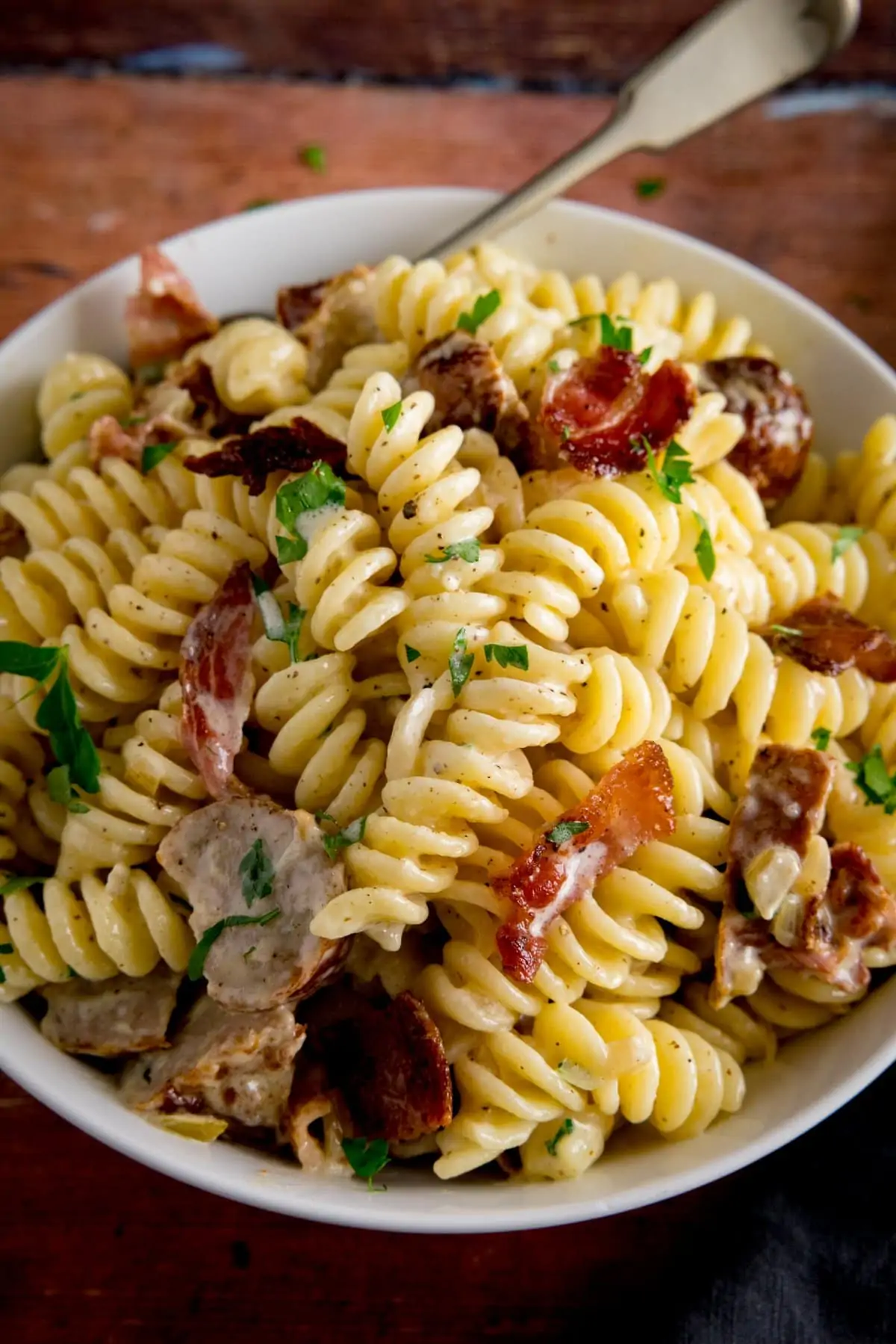 creamy pasta with sausage and bacon in a white bowl on a wooden surface. There is a fork sticking out of the pasta.