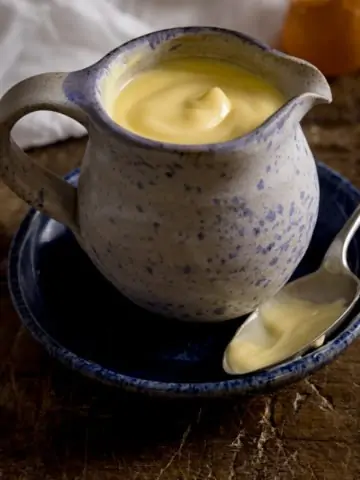 A square image of a blue jug filled with custard, sat on a blue plate with a spoon next to it. On a wooden table with a white tea towel on the side