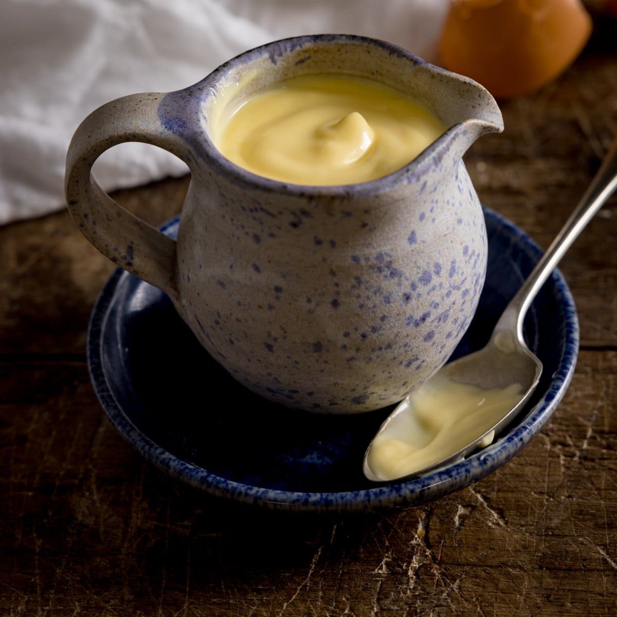 A square image of a blue jug filled with custard, sat on a blue plate with a spoon next to it. On a wooden table with a white tea towel on the side