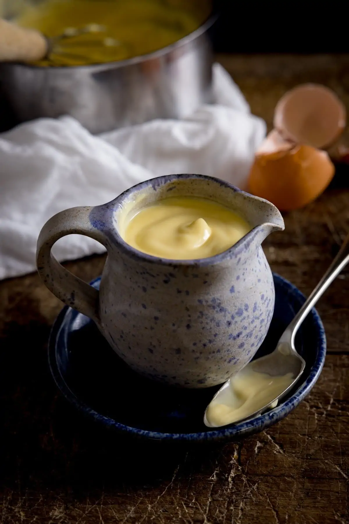 A mottled blue jug full of custard, sat on a blue plate with a spoon next to it, on a wooden table with a white tea towel in the background