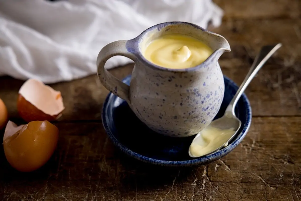 A wide image of a jug of custard on a blue plate with a spoon next to it, placed on a brown table with a white tea towel in the background
