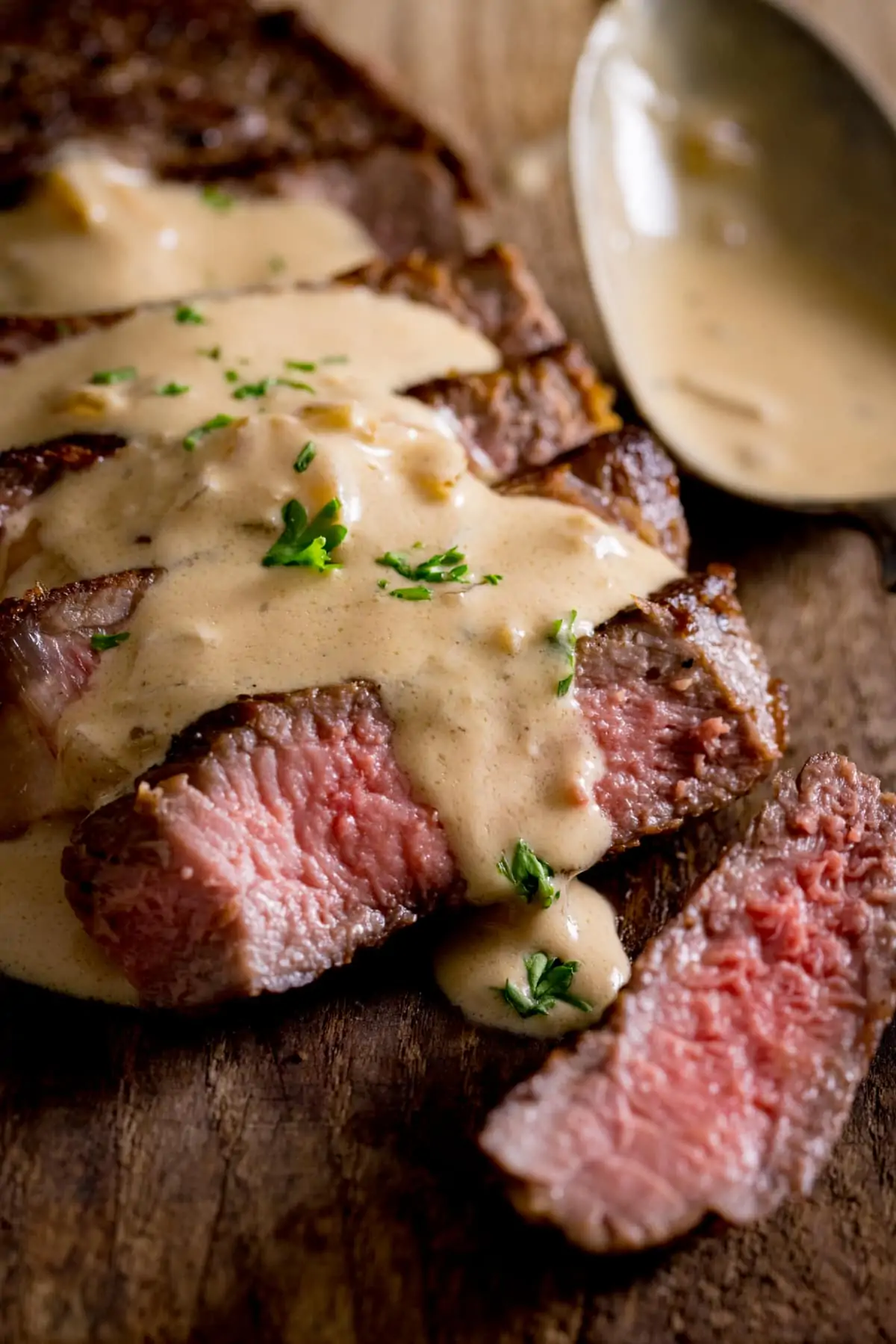 A tall image of a cooked sliced steak with diane sauce over the top with parsley sprinkled on top, a spoon is next to the steak, on a wooden board