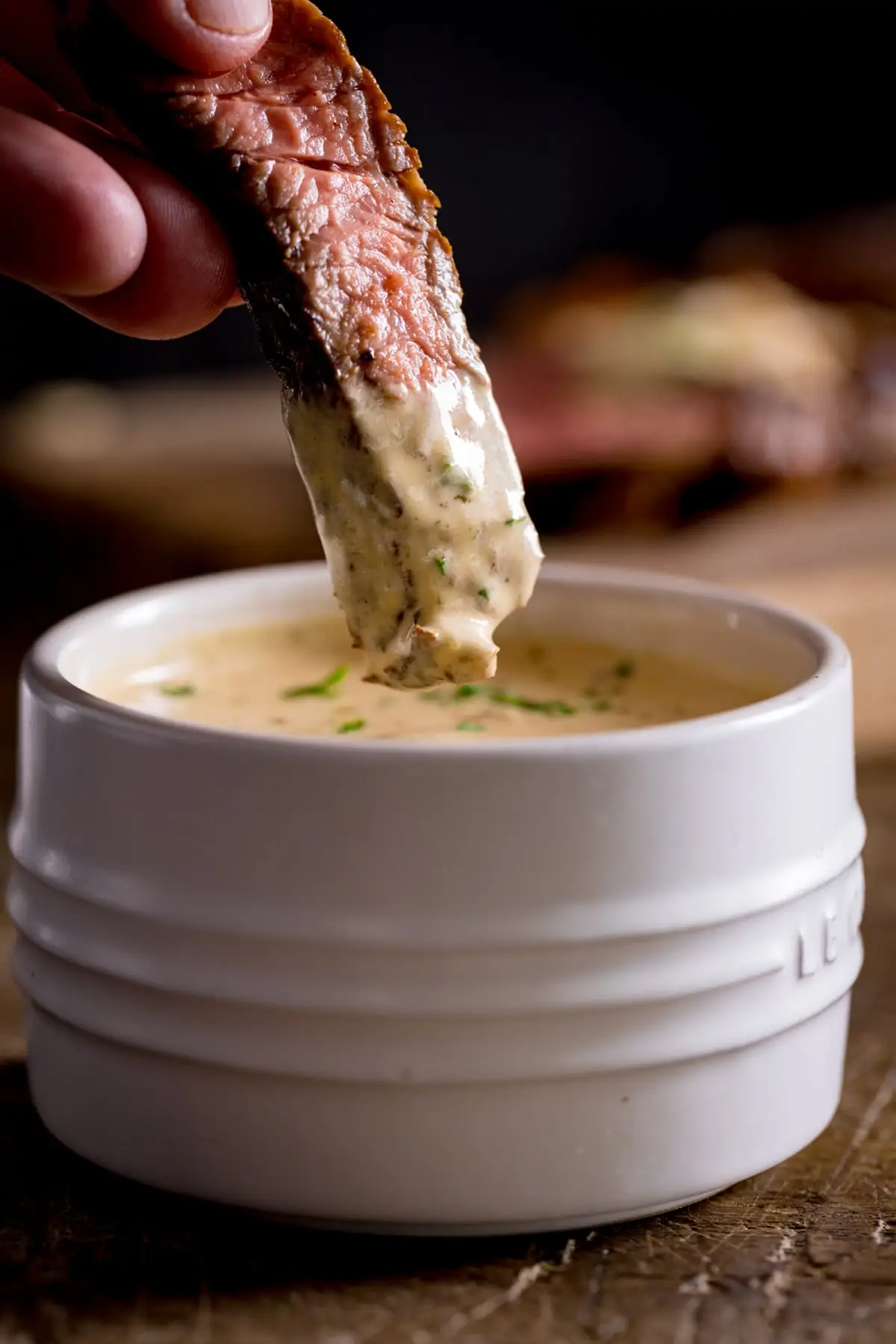 A slice of cooked steak being dipped into a white bowl of diane sauce