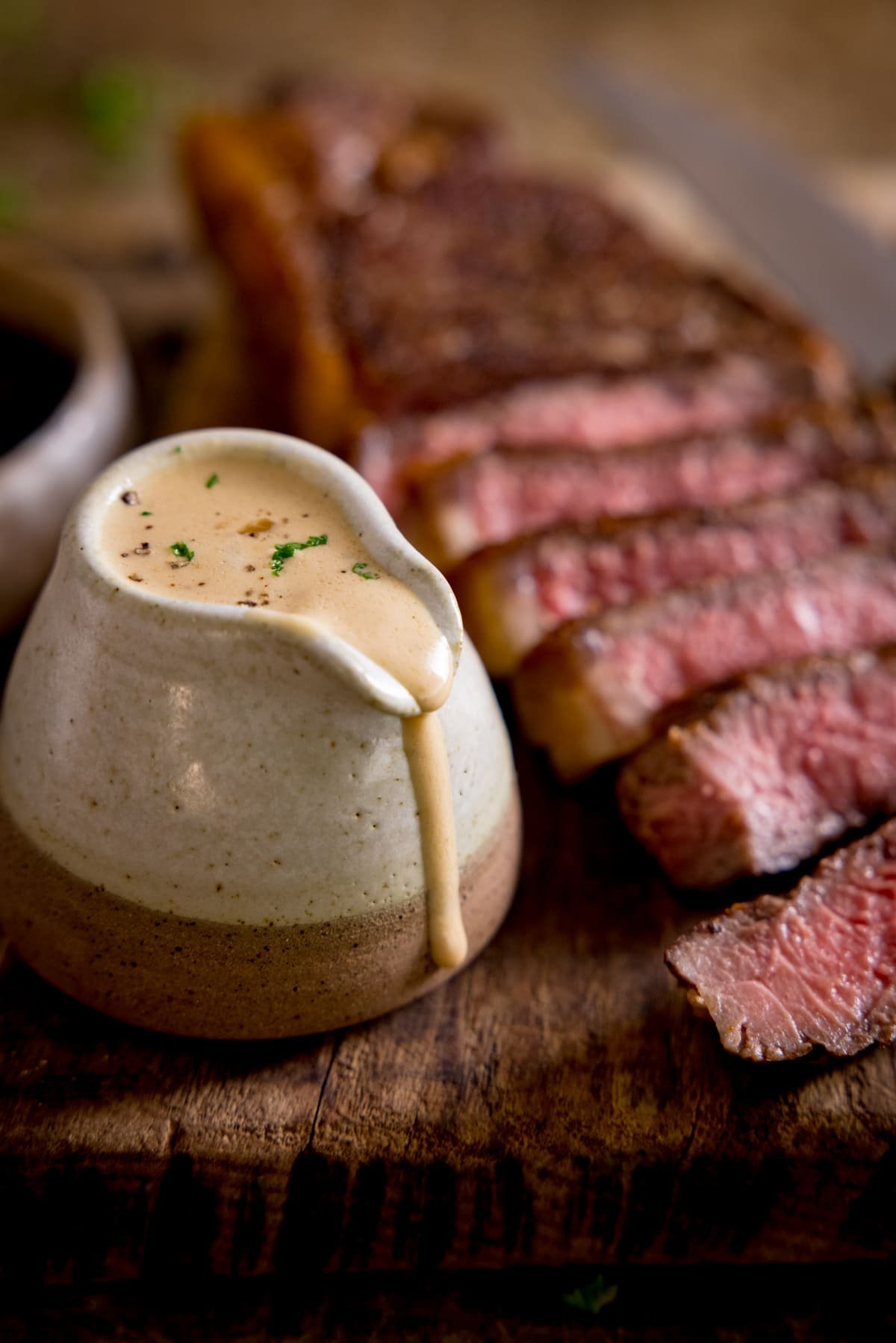 A jug of diane sauce sat next to a sliced cooked steak on a wooden board