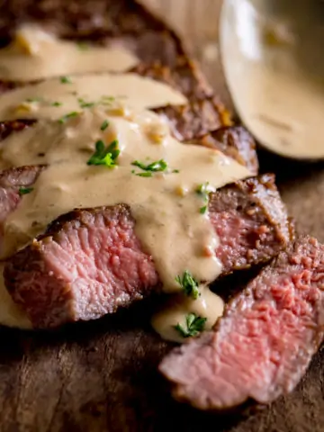 Sliced, cooked steak with diane sauce drizzled over the top on a wooden table with a silver spoon