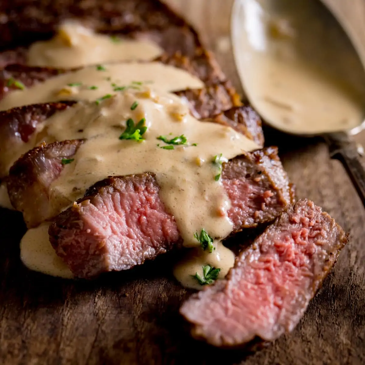 Sliced, cooked steak with diane sauce drizzled over the top on a wooden table with a silver spoon