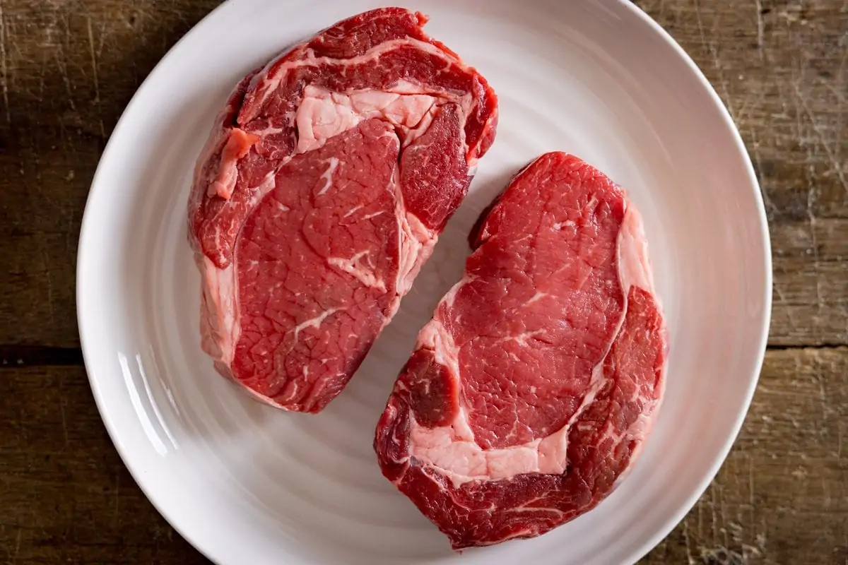 2 uncooked steaks on a white plate on a wooden table.