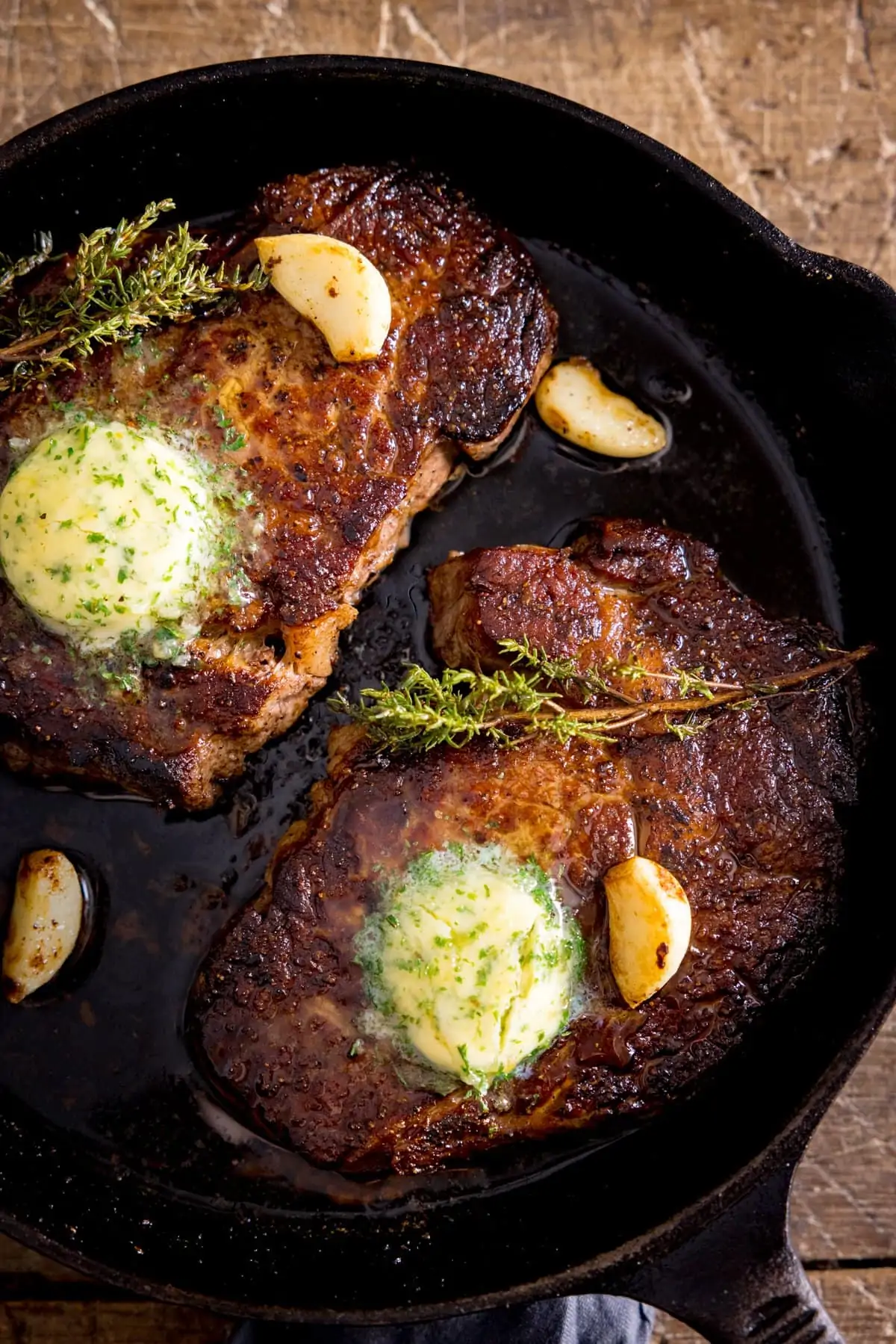 2 cooked steaks in a frying pan, topped with garlic butter, thyme and garlic