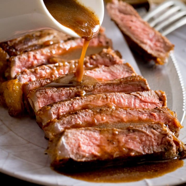 Sliced and cooked steak with red wine jus being poured over the top from a white jug, a fork with steak on it is on the side of the plate