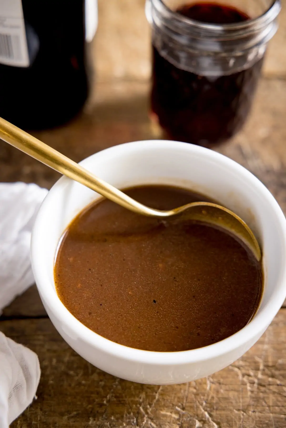 A tall image of a white bowl of red wine jus being stirred with a spoon on a wooden table, with jars in the background and a tea towel