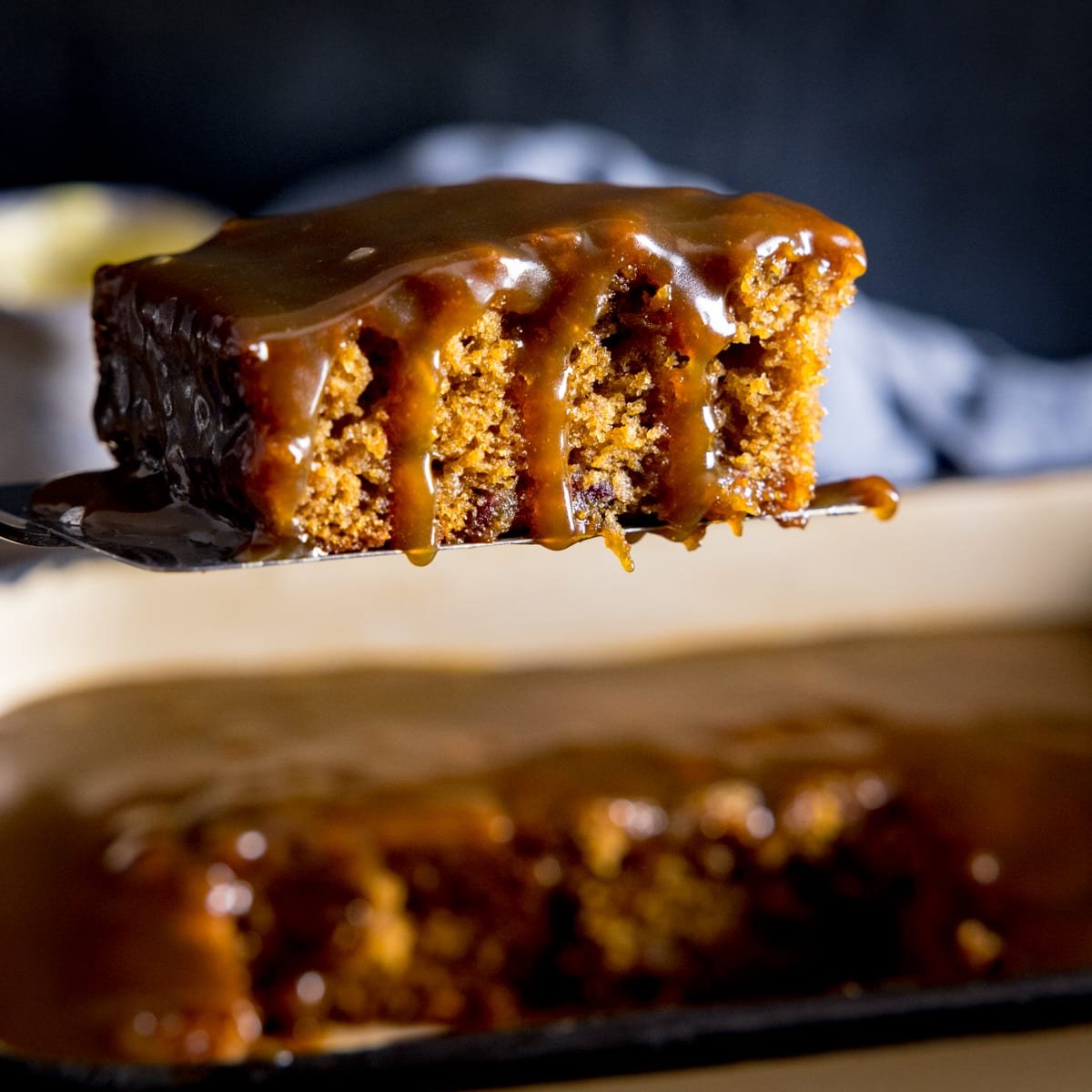 Square image of a piece of sticky toffee pudding being lifted from a baking tin
