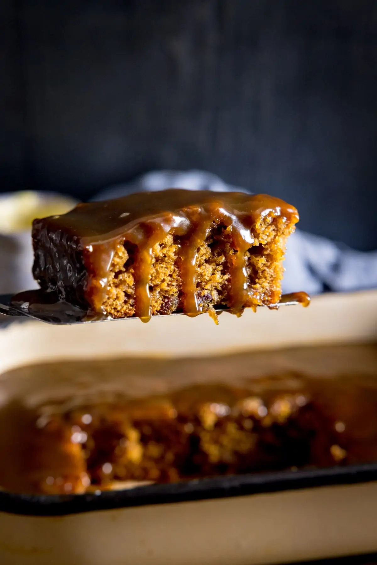 Tall image of a piece of sticky toffee pudding being lifted from a baking tray