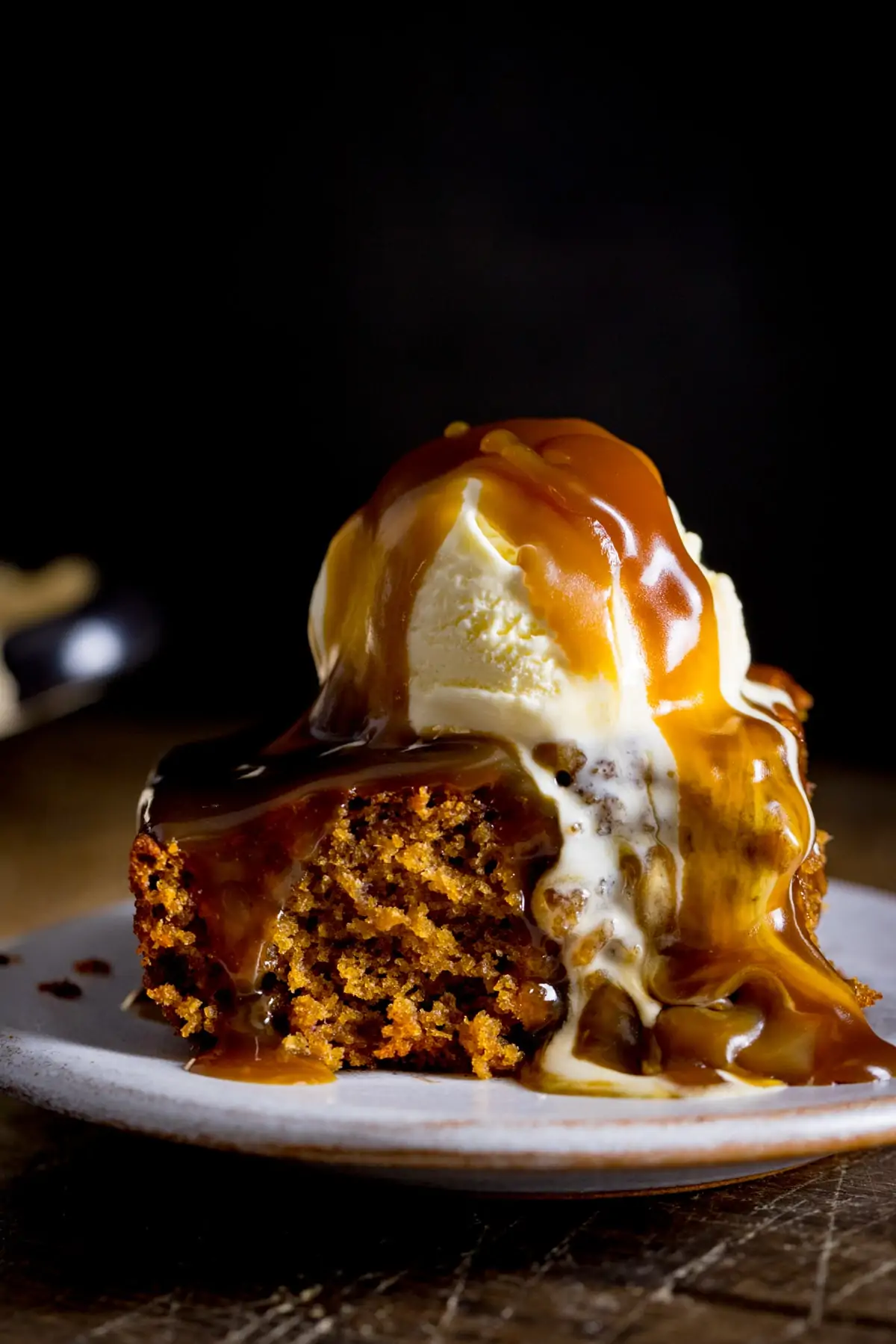 A piece of sticky toffee pudding on a white plate, topped with ice cream and toffee sauce against a dark background.