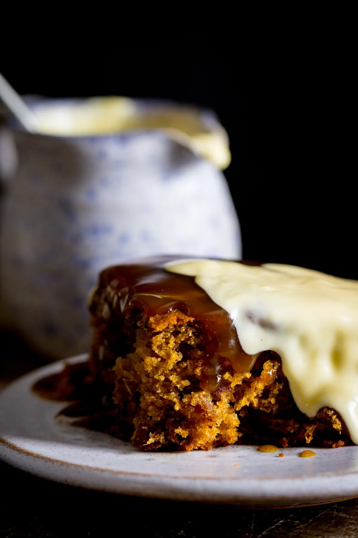Piece of sticky toffee pudding on a plate, topped with custard. Jug of custard in the background.