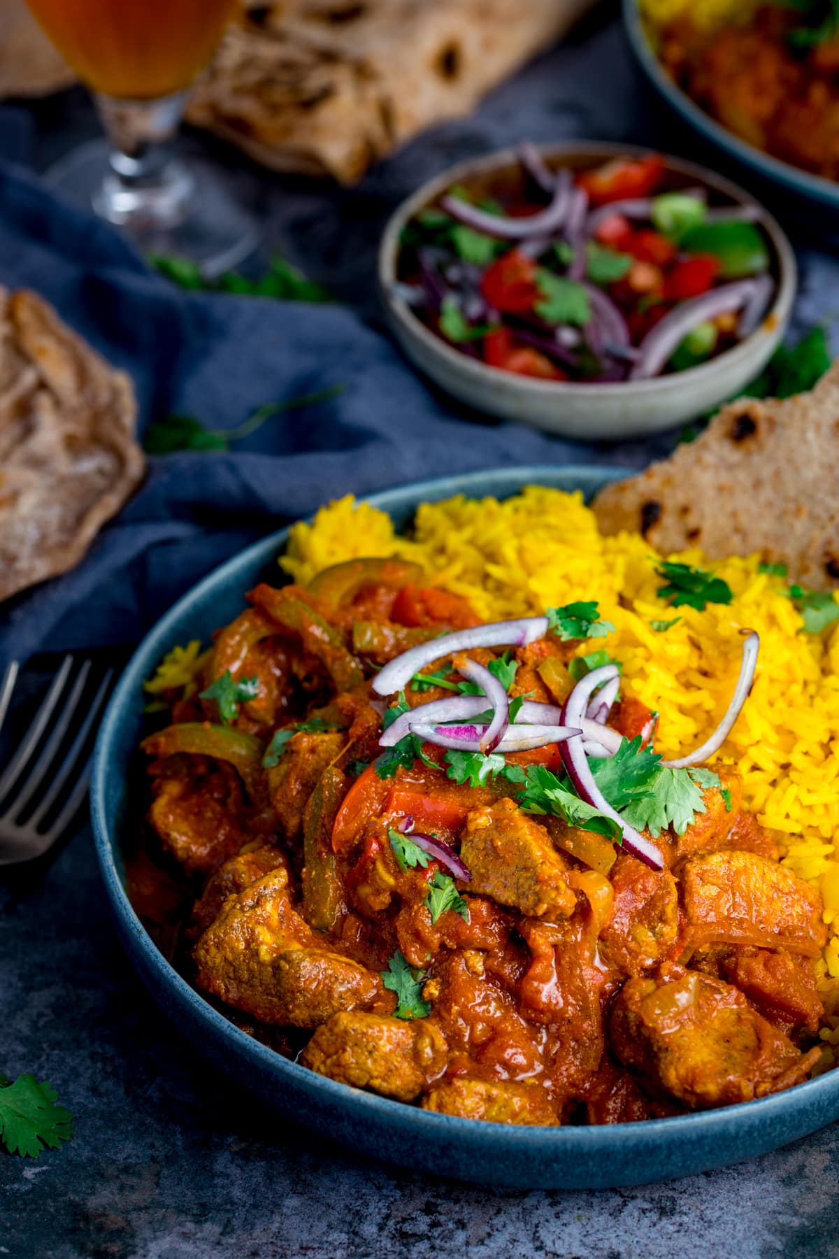 Chicken Jalfrezi with pilau rice in a blue bowl on a blue background. There is a bowl of tomato and onion salad in the background, along with forks, pieces of chapati and a further bowl of food just in shot.