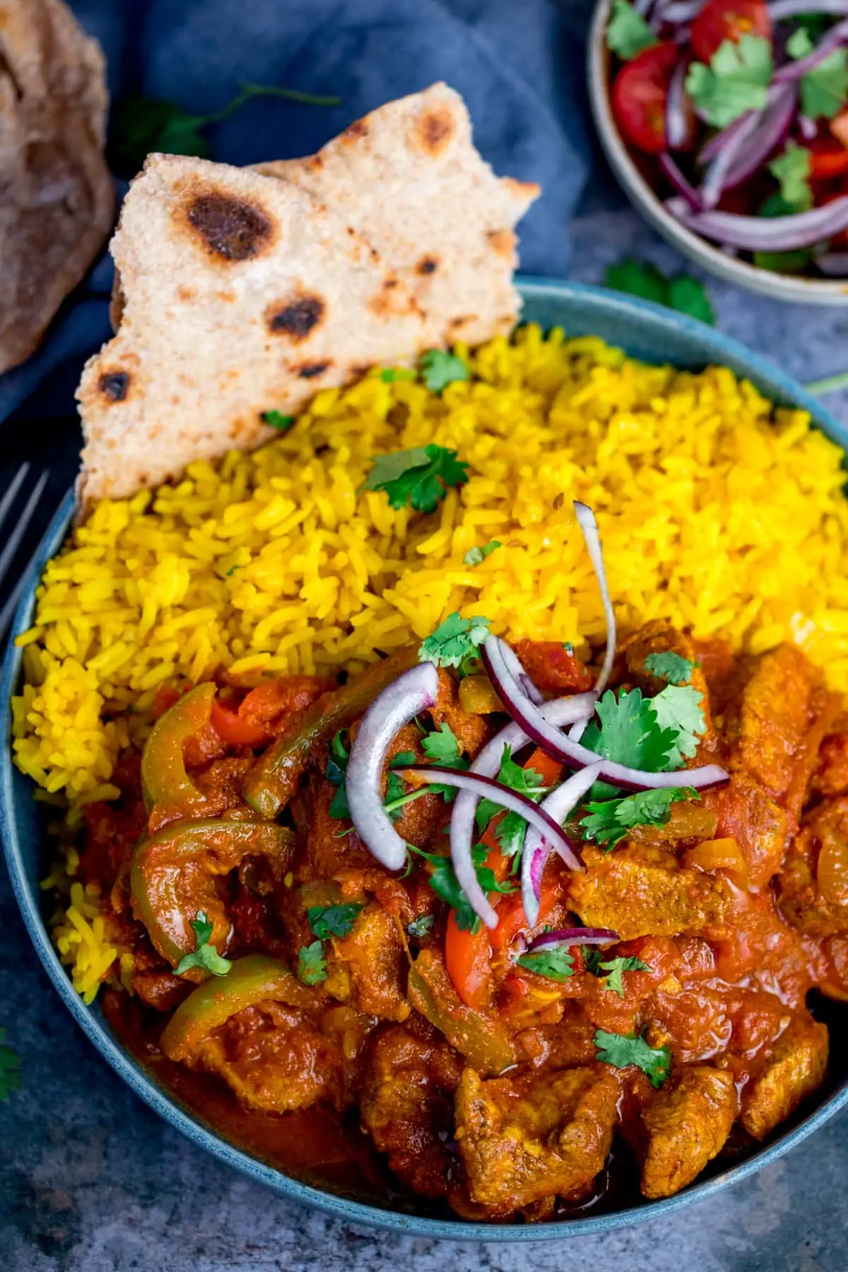 Overhead image of Chicken Jalfrezi with pilau rice in a blue bowl. The curry is topped with red onion and coriander and there is a piece of chapati in the edge of the bowl.