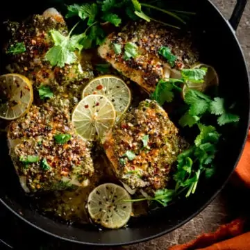 A square photo of a big pan of coriander lime cod bake, garnished with slices of lime and coriander stalks, on a table with an orange napkin to the side.