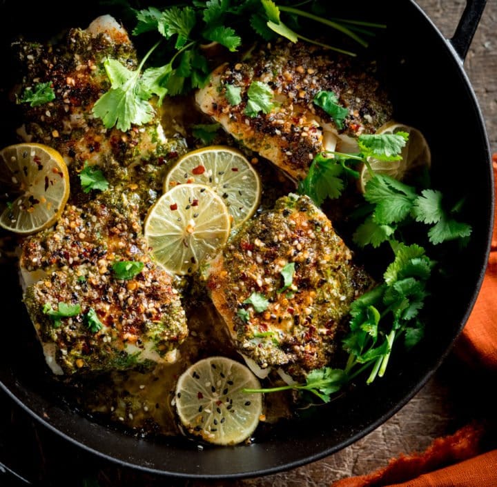 A square photo of a big pan of coriander lime cod bake, garnished with slices of lime and coriander stalks, on a table with an orange napkin to the side.