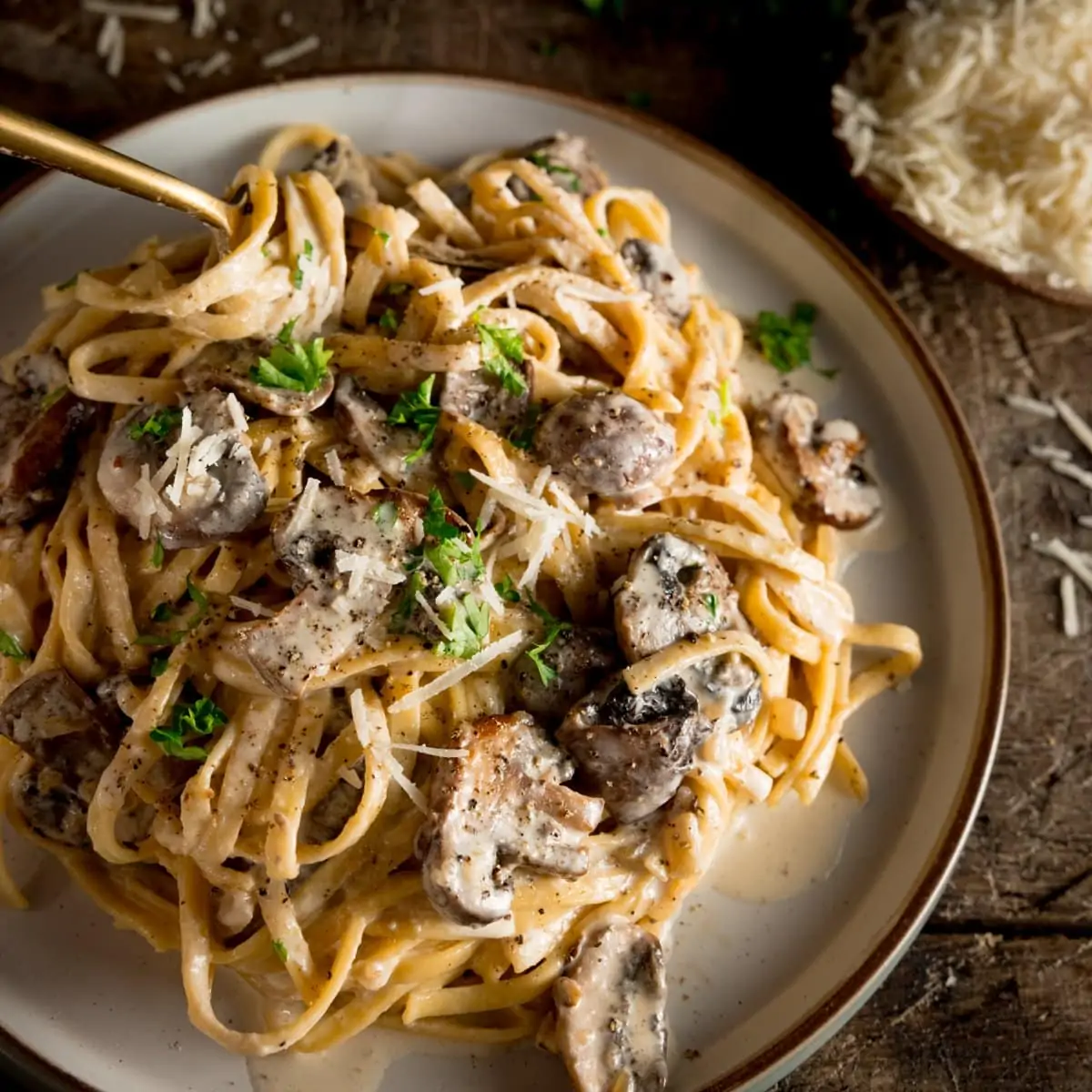 a square shot of creamy mushroom pasta on a white plate with a spoon dug in, on a wooden board with some parmesan in the background