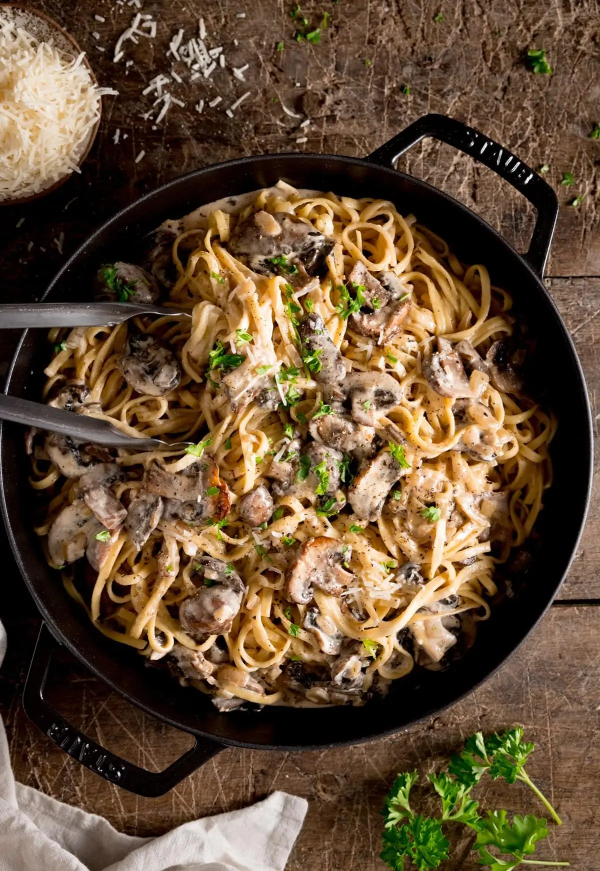 A big pan of creamy mushroom pasta with a set of metal tongs stuck in,. The pan is on a wooden background with parsley and grated parmesan scattered around.