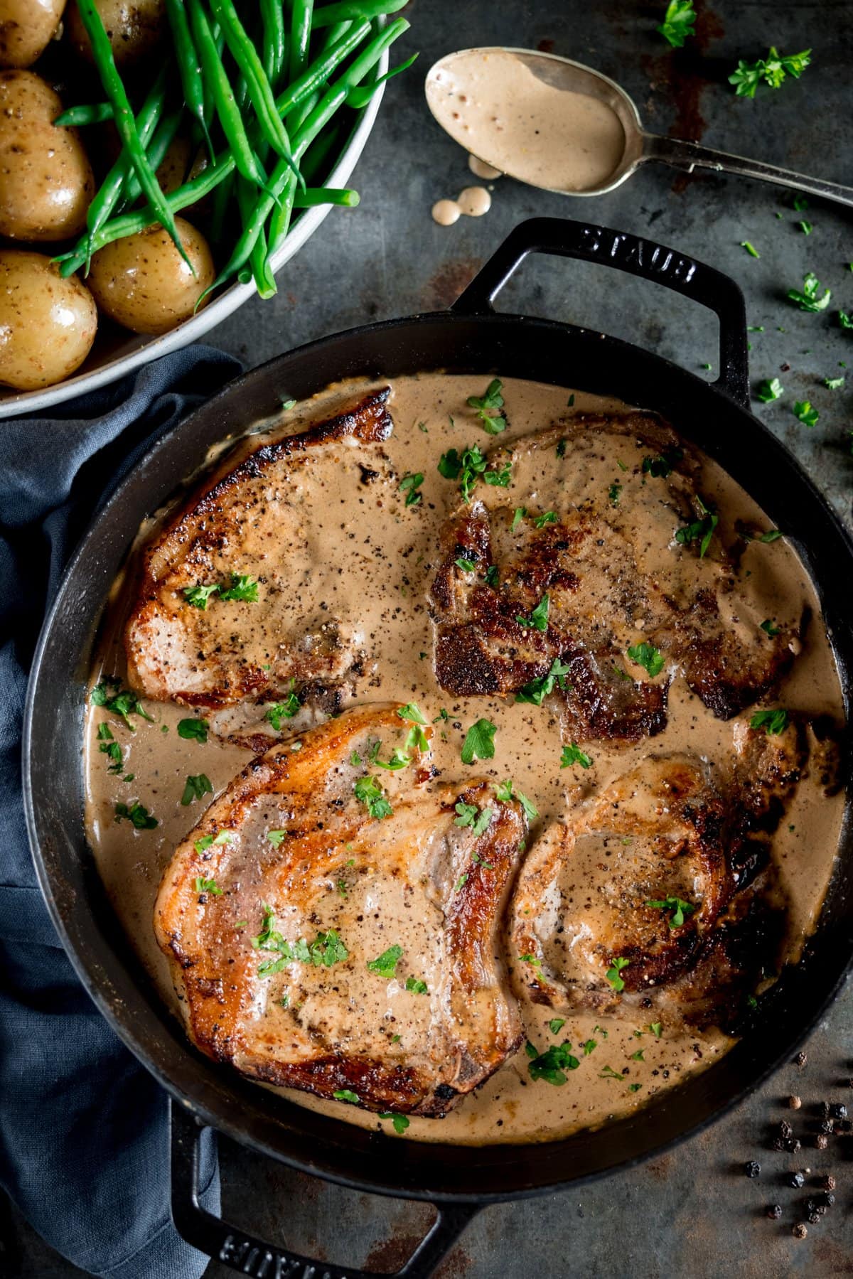 A photo looking down on a big pan of pork chops with creamy mustard sauce, and a bowl of baby new potatoes and green beans on the side, on a grey table