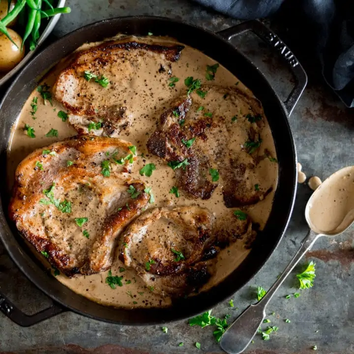 A pan of pork chops and creamy mustard sauce with a spoon laid next to it, on a grey table, a bowl of potatoes next to the pan.