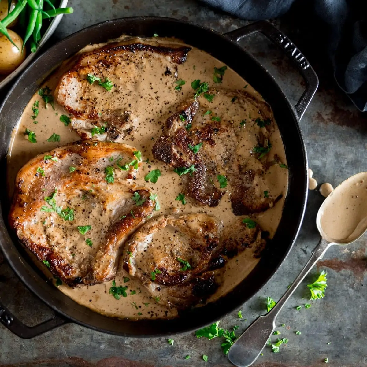A pan of pork chops and creamy mustard sauce with a spoon laid next to it, on a grey table, a bowl of potatoes next to the pan.