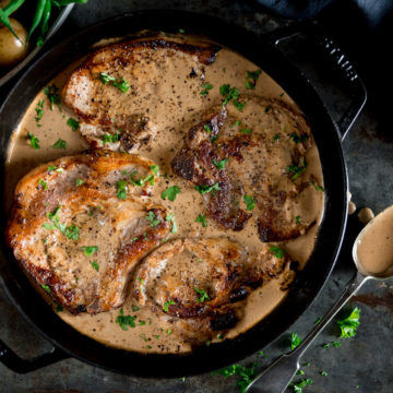 A big pot of Pork Chops with creamy mustard sauce, sprinkled with parsley. On a grey table with a spoon on the side