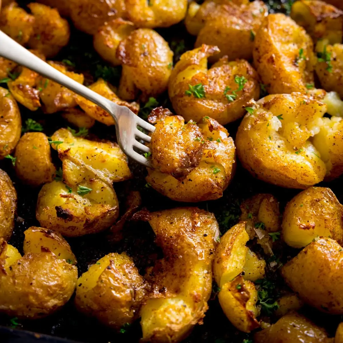 A square photo of a tray of crispy smashed potatoes, with a fork with a smashed potato on the end