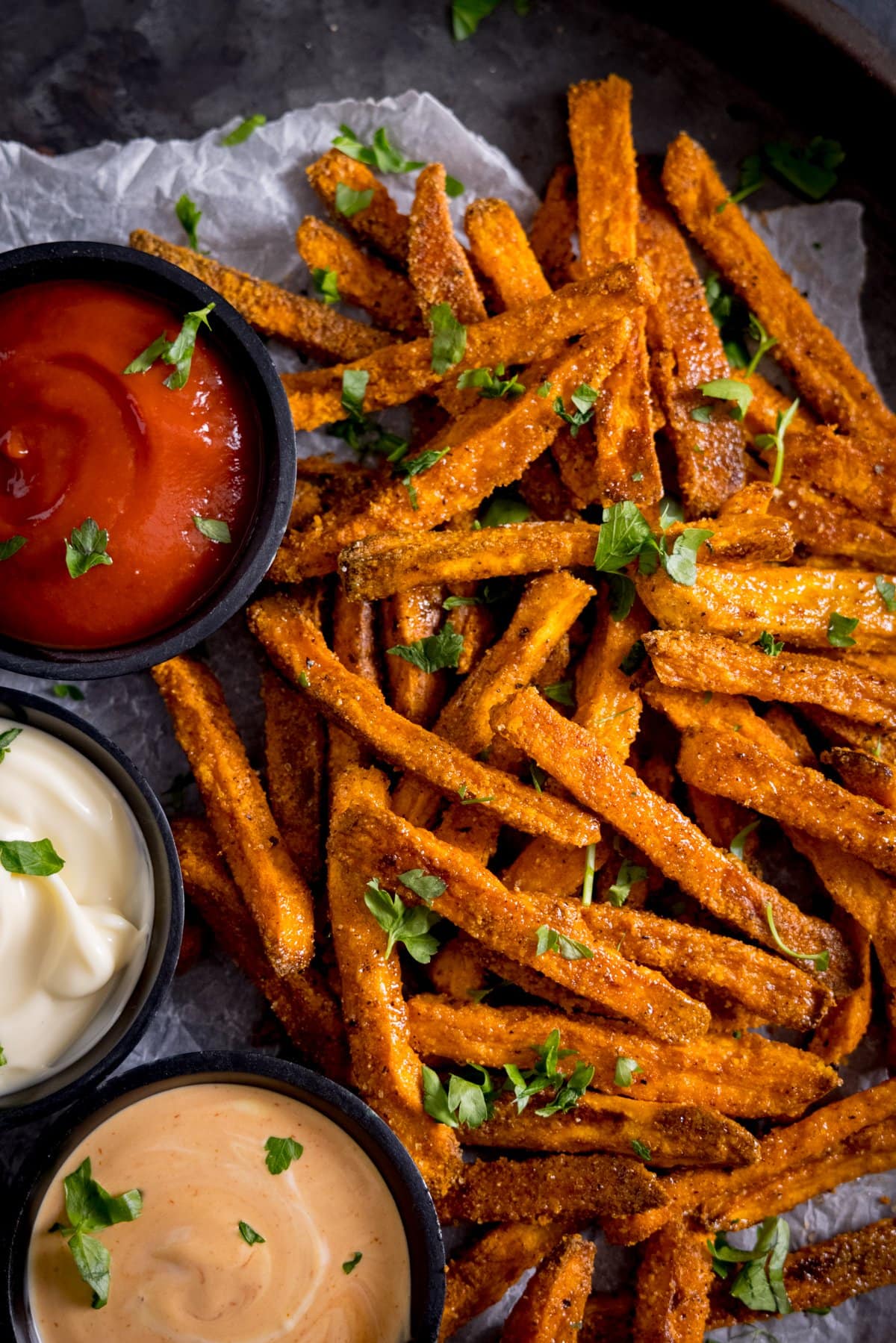 Cooked sweet potato fries sprinkled with chopped parsley with ketchup and mayo on the side in small bowls